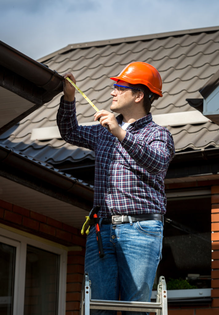 Young handyman standing on high ladder and measuring roof with tape